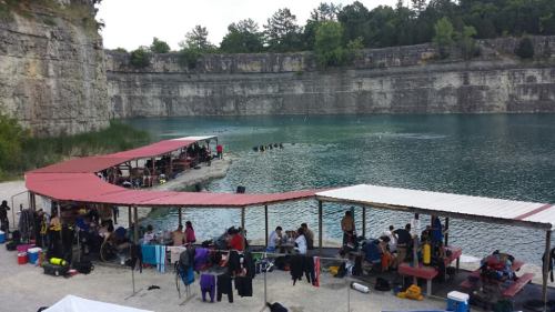 A group of people gather by a blue lake, with tents and tables set up along the shore for activities and diving.