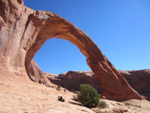 A large natural rock arch stands against a clear blue sky, with people and vegetation visible in the foreground.