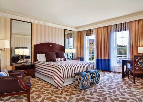 A stylish hotel room featuring a striped bed, decorative curtains, mirrors, and patterned ottomans.