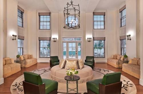 A spacious, elegantly furnished lounge with large windows, green chairs, and a central round table with flowers.