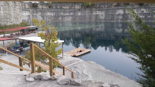 A serene quarry lake with a wooden dock, surrounded by rocky cliffs and greenery, under a cloudy sky.
