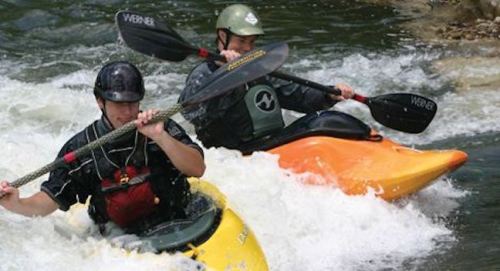 Two kayakers navigate a rapid river, one in a yellow kayak and the other in an orange kayak, both paddling vigorously.