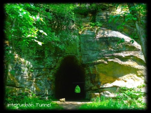 A rocky tunnel entrance surrounded by lush greenery, with sunlight illuminating the path inside.