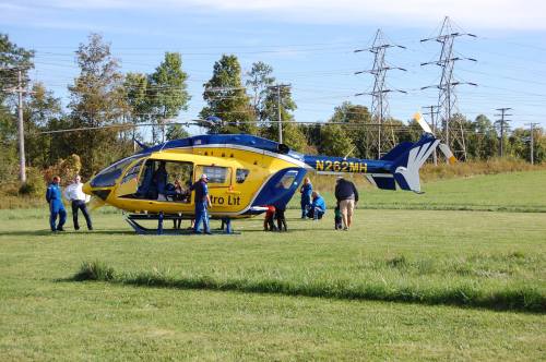 A yellow and blue helicopter on a grassy field with people preparing for a medical transport. Power lines in the background.