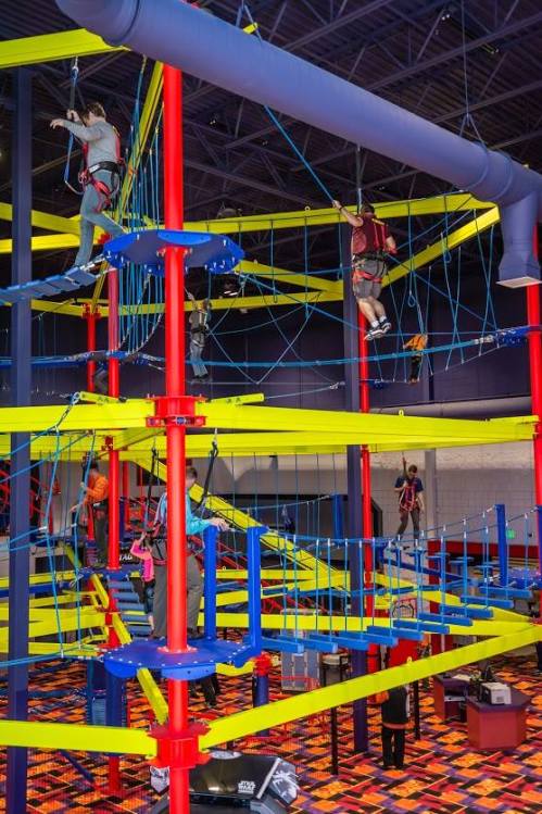 A colorful indoor climbing structure with people navigating ropes and platforms at various heights.