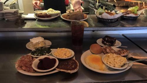 A table with various dishes including meats, sides, and desserts, with a person serving food in the background.