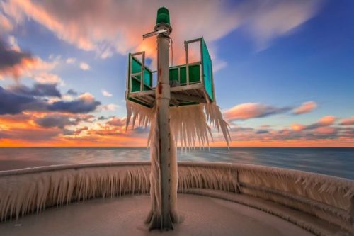A frozen lighthouse with icicles, set against a colorful sunset over the ocean.