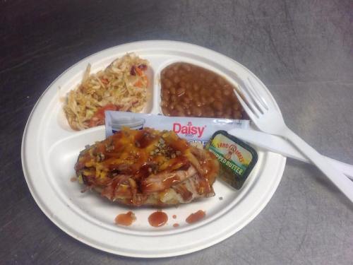 A cafeteria tray with a meat dish topped with cheese, coleslaw, baked beans, and a butter packet, accompanied by utensils.