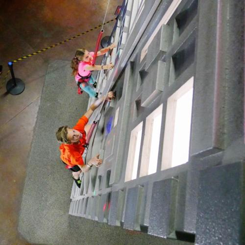 Two children climbing a vertical wall in a climbing gym, focused and determined. Safety gear is visible.