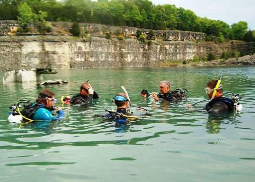 A group of six divers in wetsuits gathers in a calm, greenish water, surrounded by rocky cliffs and trees.