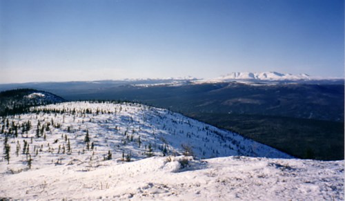 Snow-covered mountains and hills under a clear blue sky, with a vast landscape stretching into the distance.