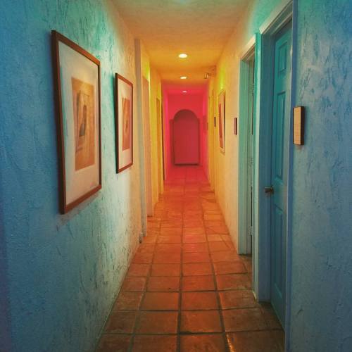A brightly lit hallway with colorful lighting, framed artwork on the walls, and doors on either side.