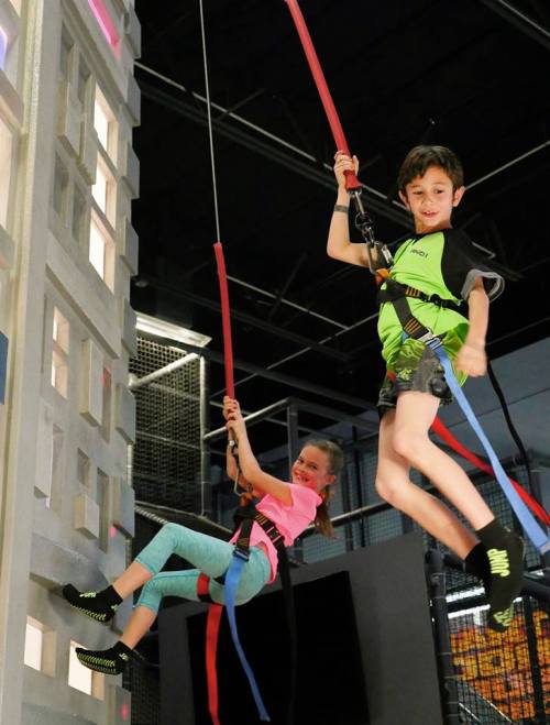 Two children in harnesses are climbing a wall, one swinging down while the other hangs on, both smiling and having fun.