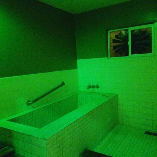 A dimly lit bathroom with green lighting, featuring a tiled bathtub and a window with stained glass.
