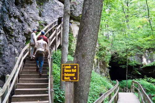 Hikers ascend wooden stairs in a lush forest, with a sign indicating nearby attractions like Balanced Rock and Dance Hall.