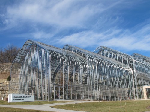 A modern glass greenhouse with a curved design, set against a blue sky and grassy landscape.