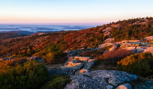 A scenic view of colorful autumn foliage on rocky terrain, overlooking a calm body of water and distant islands at sunset.