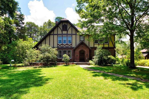 A large, charming two-story house with a mix of brick and stucco, surrounded by lush greenery and a well-maintained lawn.