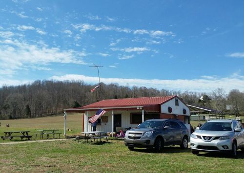 A small building with a red roof, American flags, and parked cars in a grassy area under a blue sky with clouds.