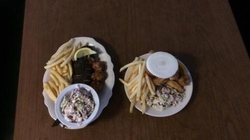 Two plates of food on a wooden table: one with fries, fried shrimp, and coleslaw; the other with fries and a side of greens.