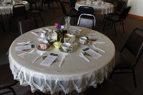 A round table set for tea with floral tableware, utensils, and menus, surrounded by chairs in a cozy room.