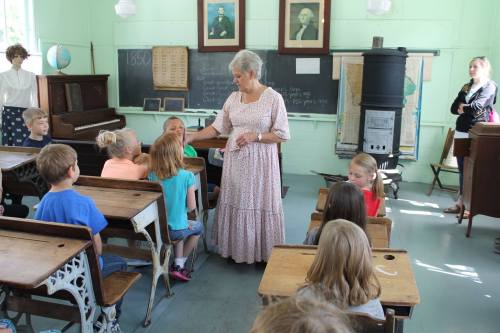 A teacher in a vintage classroom engages with a group of children seated at old-fashioned desks.