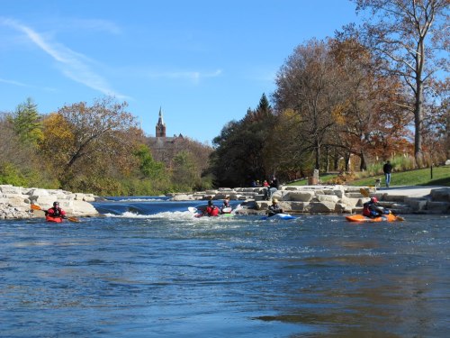 A scenic river with kayakers navigating the water, surrounded by trees and a distant building under a clear blue sky.