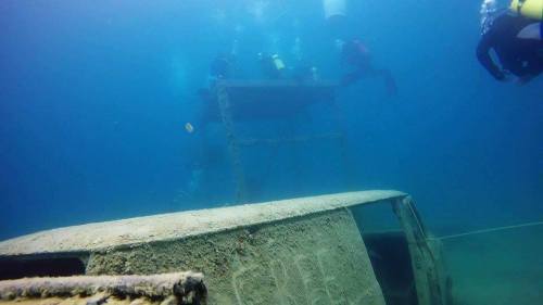 Divers explore a submerged vehicle and a structure in clear blue water, surrounded by marine life.