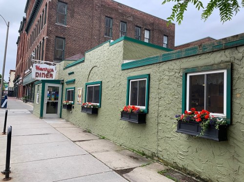 A green diner with flower boxes under the windows, located on a city street with brick buildings in the background.