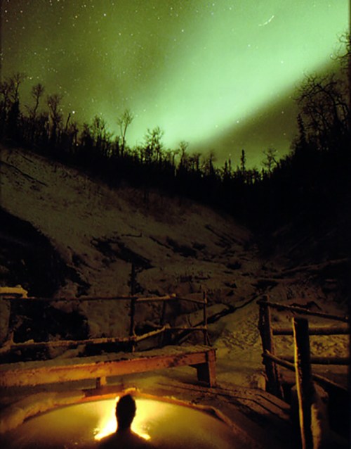 A person sits in a hot spring, surrounded by snow, under a vibrant display of the northern lights.