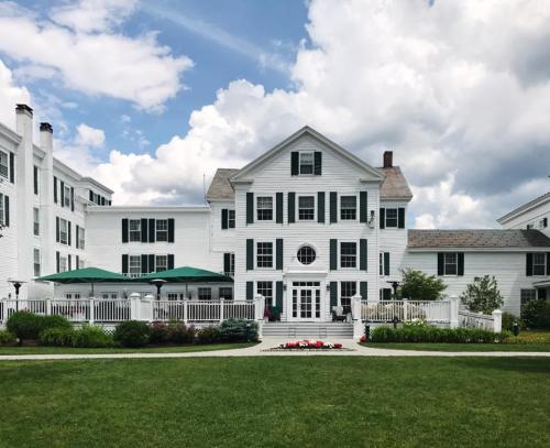A large white house with green shutters, surrounded by gardens and a patio, under a partly cloudy sky.