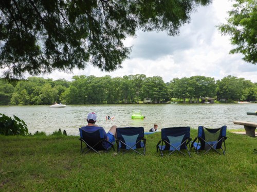A person sits in a chair by a lake, watching others swim and enjoy the water on a cloudy day.