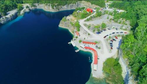 Aerial view of a serene blue lake surrounded by greenery, with a parking area and buildings along the shore.