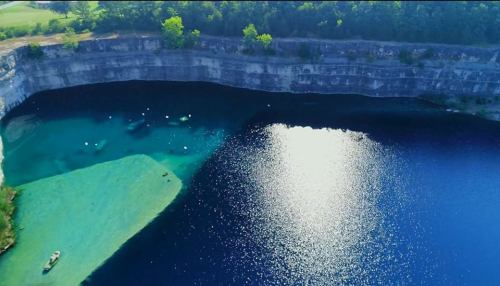 Aerial view of a deep blue lake surrounded by rocky cliffs and lush greenery, with small boats on the water.