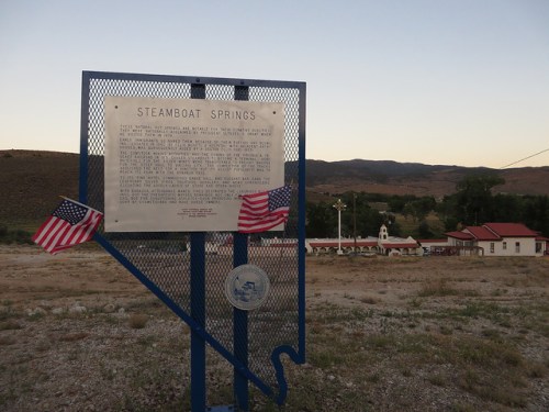 A historical sign about Steamboat Springs, with American flags, set against a backdrop of mountains and a building.