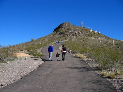 A family walks along a winding path towards a hill under a clear blue sky, with a few people visible in the distance.