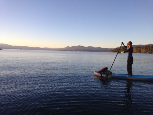 A person paddleboarding on calm water during sunset, with mountains in the background and a clear sky.