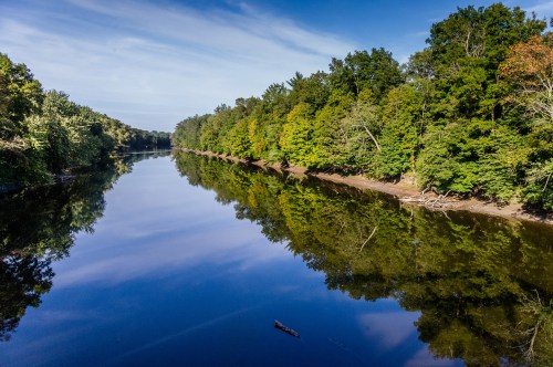 A serene river reflects lush green trees under a clear blue sky, creating a peaceful natural landscape.