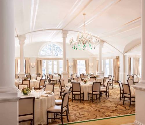 Elegant dining room with round tables, white tablecloths, and a chandelier, featuring large windows and soft lighting.