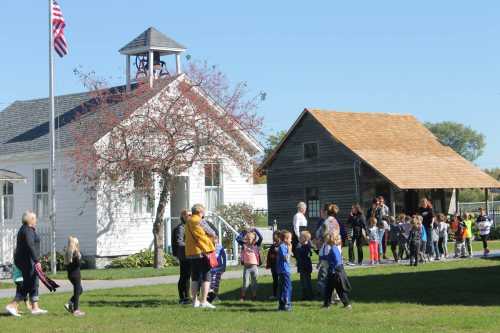 A group of children and adults gather outside a white building and a wooden structure on a sunny day.