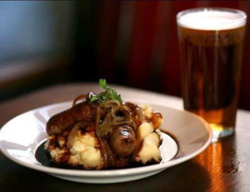 A plate of sausages on mashed potatoes, topped with gravy and onions, alongside a pint of beer.