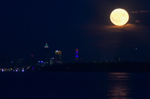 A bright full moon illuminates the night sky over a city skyline with colorful lights reflecting on the water.