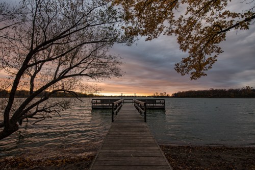 A wooden dock extends into a calm lake at sunset, framed by trees and a colorful sky.