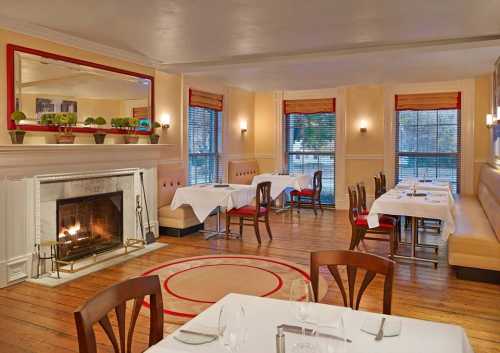 Cozy restaurant interior with a fireplace, wooden floors, and neatly set tables by large windows.