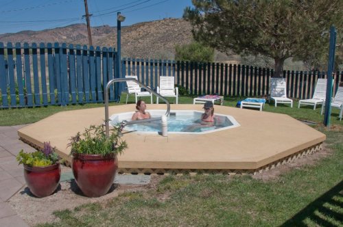 Two people relax in a hot tub surrounded by greenery and lounge chairs, with a blue fence and mountains in the background.