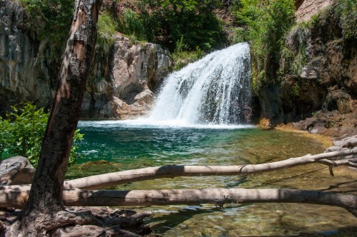 A serene waterfall cascades into a clear pool, surrounded by lush greenery and rocky cliffs.