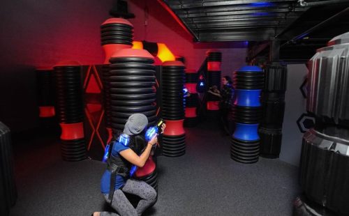 A person crouches with a laser tag gun in a colorful arena filled with obstacles and dim lighting.