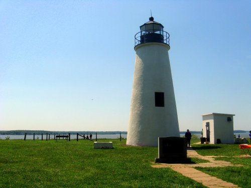A tall white lighthouse stands on green grass by the water, with a clear blue sky in the background.
