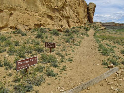 A dirt path leads to the Petroglyph Trail, flanked by signs and rocky cliffs under a blue sky.