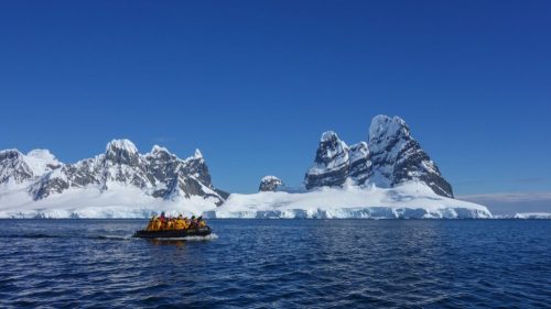 A small boat with passengers navigates icy waters near towering snow-covered mountains under a clear blue sky.